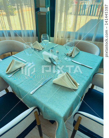 Dining table is set with cutlery and glassware in a cafe during the daytime near a beach area with sunlight streaming through the windows 141445397