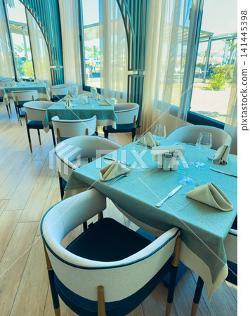 Dining area with tables set for lunch in a restaurant with large windows and views of the outdoors during daylight hours Dining area with tables set for lunch in a restaurant with large windows and views of the outdoors during daylight hours 141445398