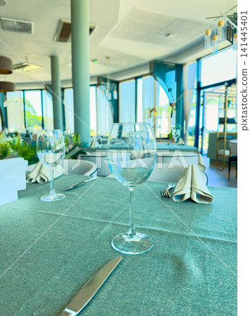 Restaurant table set with glasses and cutlery in a bright dining area near the ocean during daytime Restaurant table set with glasses and cutlery in a bright dining area near the ocean during daytime 141445401