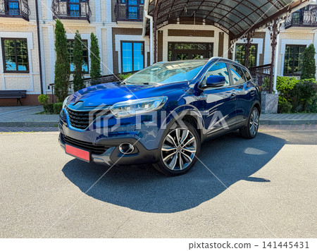 Blue SUV parked outside a large building with dark roof, green trees, and blue windows on a sunny day in the city Blue SUV parked outside a large building with dark roof, green trees, and blue windows on a sunny day in the city 141445431