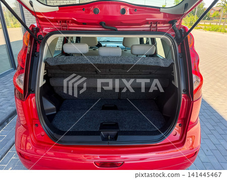 Car trunk space shows a clean area with flat seats in a red vehicle parked beside a building on a sunny day with some greenery 141445467