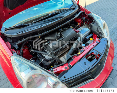Engine of a red car is open with hood lifted showing internal parts in a garage on a sunny day Engine of a red car is open with hood lifted showing internal parts in a garage on a sunny day 141445473