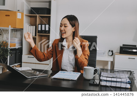 Young Asian professional business woman calculating finances and analyzing data charts on computer, smiling while working on accounting project in a modern office, planning budget Young Asian professional business woman calculating finances and analyzing data charts on computer, smiling while working on accounting project in a modern office, planning budget 141445876