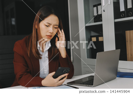 Smiling asian businesswoman talking on phone and using laptop while working at office desk Smiling asian businesswoman talking on phone and using laptop while working at office desk 141445904
