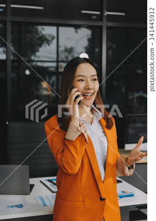 Smiling asian businesswoman talking on phone and using laptop while working at office desk Smiling asian businesswoman talking on phone and using laptop while working at office desk 141445912
