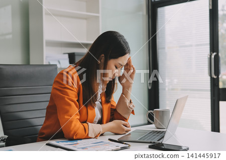 Office Discomfort: A female office worker in a red shirt experiences neck pain while working on her laptop, surrounded by office supplies and a calculator, indicating a stressful work environment. Office Discomfort: A female office worker in a red shirt experiences neck pain while working on her laptop, surrounded by office supplies and a calculator, indicating a stressful work environment. 141445917