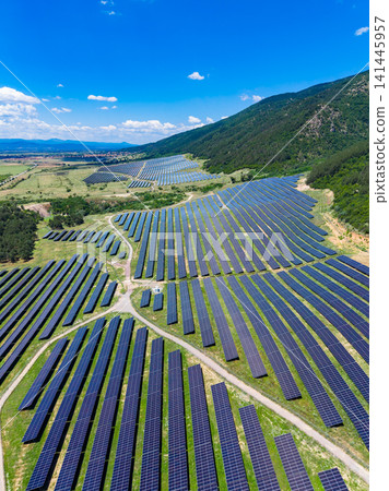 Solar panels cover large fields under a blue sky with scattered clouds in a rural area near mountains on an sunny day Solar panels cover large fields under a blue sky with scattered clouds in a rural area near mountains on an sunny day 141445957