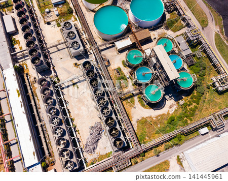 Industrial facility with large tanks, cooling systems, and pipes located near a water body during daylight hours Industrial facility with large tanks, cooling systems, and pipes located near a water body during daylight hours 141445961