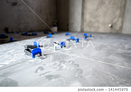 Grey floor tiles installing in a new house or apartment, showing clips and wedges of a tile leveling system ensuring an even surface during the renovation process Grey floor tiles installing in a new house or apartment, showing clips and wedges of a tile leveling system ensuring an even surface during the renovation process 141445978
