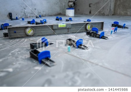 Tiler installing new ceramic floor tiles with a tile leveling system, ensuring even surfaces for house renovation work on a construction site Tiler installing new ceramic floor tiles with a tile leveling system, ensuring even surfaces for house renovation work on a construction site 141445986