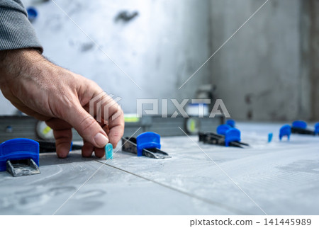 Hand of a tiler installing new gray ceramic floor tiles on adhesive mortar, using a tile leveling system to ensure a perfectly even and flat surface during home renovation Hand of a tiler installing new gray ceramic floor tiles on adhesive mortar, using a tile leveling system to ensure a perfectly even and flat surface during home renovation 141445989