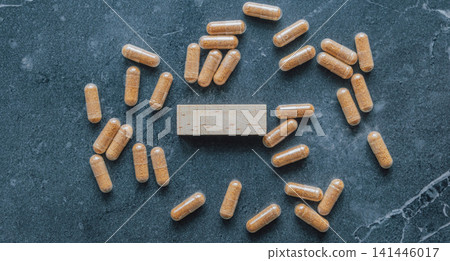 Top view flat lay of herbal brown pill capsules scattered around blank wooden block on dark stone surface for natural dietary supplement health concept Top view flat lay of herbal brown pill capsules scattered around blank wooden block on dark stone surface for natural dietary supplement health concept 141446017