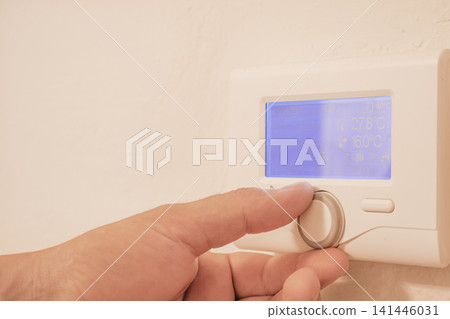 Close up view of mans hand adjusting digital thermostat controller dial on wall for home heating temperature efficiency and energy saving utility concept Close up view of mans hand adjusting digital thermostat controller dial on wall for home heating temperature efficiency and energy saving utility concept 141446031