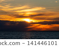 Dramatic golden sunset over calm sea with dark clouds cutting through sun. Beautiful warm light reflecting on water surface. Hope and serenity concept. 141446101