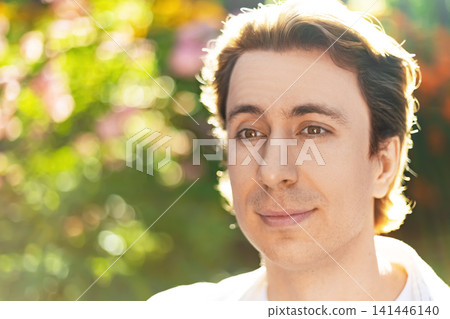 Close-up portrait of a handsome young man with a calm smile. Outdoor nature setting with sunny backlit bokeh. Peaceful male face looking away. Close-up portrait of a handsome young man with a calm smile. Outdoor nature setting with sunny backlit bokeh. Peaceful male face looking away. 141446140