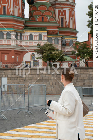Woman looks toward Saint Basils Cathedral in central Moscow while holding smartphone on cloudy summer day. Urban lifestyle, travel, sightseeing, architecture, cultural heritage, city exploration Woman looks toward Saint Basils Cathedral in central Moscow while holding smartphone on cloudy summer day. Urban lifestyle, travel, sightseeing, architecture, cultural heritage, city exploration 141446429