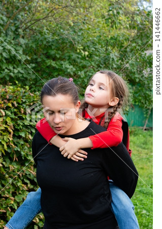 A mother gives her daughter a piggyback ride through the yard, showcasing a heartwarming moment of family bonding and simple joy. A mother gives her daughter a piggyback ride through the yard, showcasing a heartwarming moment of family bonding and simple joy. 141446662