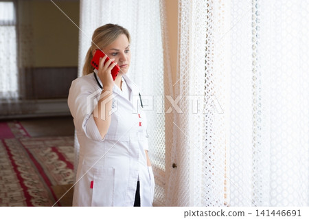 Female doctor on phone looking out window. Medical professional wearing white coat. Healthcare professional. Telemedicine concept. Patient care. Female doctor on phone looking out window. Medical professional wearing white coat. Healthcare professional. Telemedicine concept. Patient care. 141446691