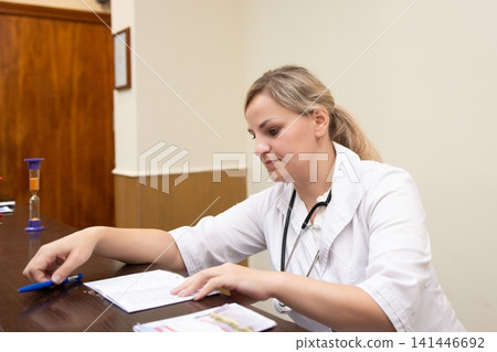 Professional woman in a white coat reviewing paperwork at a desk. She looks focused, wearing a stethoscope and holding a blue pen. Professional woman in a white coat reviewing paperwork at a desk. She looks focused, wearing a stethoscope and holding a blue pen. 141446692