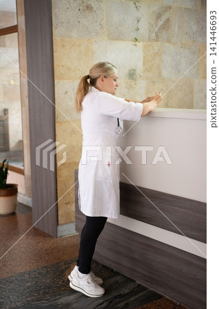 Healthcare worker in uniform checking her phone at a reception desk, standing against stone wall, wearing black leggings and white shoes. Healthcare worker in uniform checking her phone at a reception desk, standing against stone wall, wearing black leggings and white shoes. 141446693