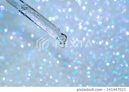 Glass dropper releasing a clear serum droplet against a sparkling blue bokeh background Glass dropper releasing a clear serum droplet against a sparkling blue bokeh background 141447021