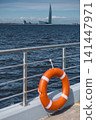An orange-colored lifebuoy hangs on the embankment fence in sunny weather, finland gulf, with the sea and a skyscraper in the background. 141447971