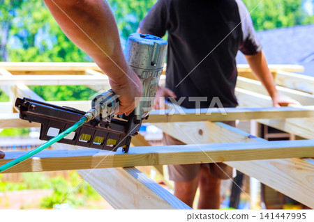 Construction workers use air nail gun to secure wooden beams in an outdoor setting Construction workers use air nail gun to secure wooden beams in an outdoor setting 141447995