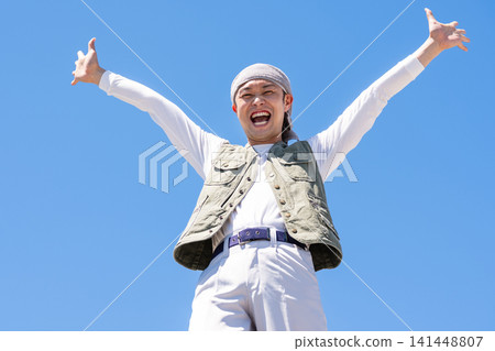 A man in work clothes raises his arms in a cheer against a blue sky. A man in work clothes raises his arms in a cheer against a blue sky. 141448807