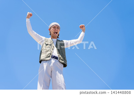 A man in work clothes strikes a victory pose against a blue sky. A man in work clothes strikes a victory pose against a blue sky. 141448808