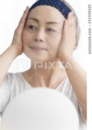 A senior woman uses a facial steamer to perform a self-care facial. A senior woman uses a facial steamer to perform a self-care facial. 141449105