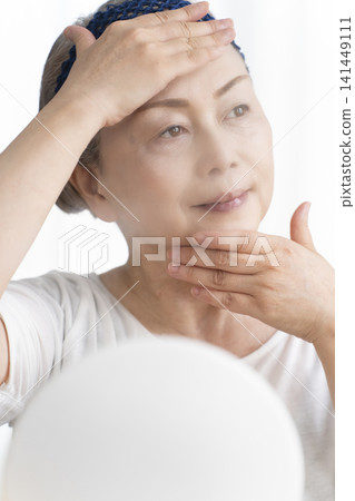 A senior woman uses a facial steamer to perform a self-care facial. 141449111