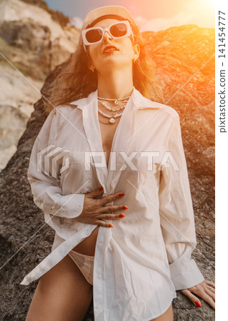 Woman summer beach posing in white shirt and sunglasses on rocky shore at sunset during vacation with copy space 141454777
