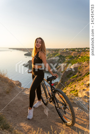 Woman mountain bike sunset posing on a high cliff overlooking the ocean during golden hour for healthy outdoor adventure and fitness 141454783