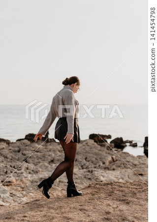 Woman ocean seashore stroll along a rocky coast wearing knitwear and a black skirt against a clear sky Woman ocean seashore stroll along a rocky coast wearing knitwear and a black skirt against a clear sky 141454798