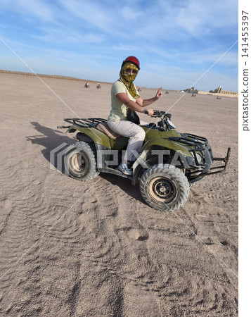 Adventurous woman casual summer outfit confidently rides green quad bike across golden desert dunes, arms raised in celebration, under a bright blue sky dynamic freedom, thrill outdoor exploration. Adventurous woman casual summer outfit confidently rides green quad bike across golden desert dunes, arms raised in celebration, under a bright blue sky dynamic freedom, thrill outdoor exploration. 141455397