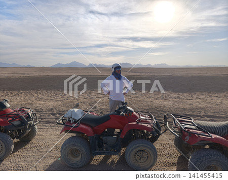 Triumphant man white outfit and protective helmet stands atop vibrant red quad bike vast desert arms raised celebration capturing pure thrill adventure freedom exploring landscapes under bright sky Triumphant man white outfit and protective helmet stands atop vibrant red quad bike vast desert arms raised celebration capturing pure thrill adventure freedom exploring landscapes under bright sky 141455415