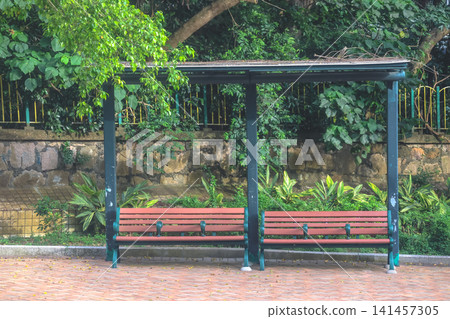Hong Kong. July 19 2026 Wooden park benches under shelter in lush garden. Hong Kong. July 19 2026 Wooden park benches under shelter in lush garden. 141457305