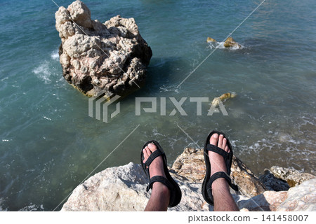Hiker feet in sandals resting on coastal cliffs overlooking Petra tou Romiou, Cyprus 141458007