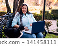 Beautiful Latina adult executive woman working in the yard of her house, with her laptop, being teased and interrupted by her cat, with a lot of humor and joy. teleworking, marketing. 141458111