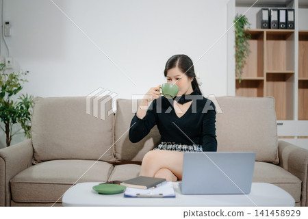 Young Asian businesswoman working remotely from home with laptop and coffee. Modern home office, freelance lifestyle, online business, productivity 141458929