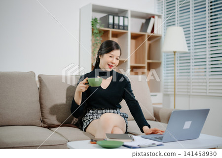 Young Asian businesswoman working remotely from home with laptop and coffee. Modern home office, freelance lifestyle, online business, productivity 141458930
