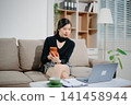 Young Asian businesswoman working remotely from home with laptop and coffee. Modern home office, freelance lifestyle, online business, productivity 141458944