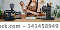 Focused Asian female barista taking stock and managing inventory with a clipboard at a modern cafe counter. 141458949