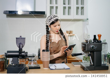 Focused Asian female barista taking stock and managing inventory with a clipboard at a modern cafe counter. Focused Asian female barista taking stock and managing inventory with a clipboard at a modern cafe counter. 141458951
