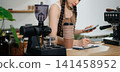 Focused Asian female barista taking stock and managing inventory with a clipboard at a modern cafe counter. 141458952
