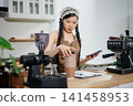 Young Asian female barista preparing fresh roasted coffee beans in a modern cafe kitchen with professional espresso equipment. 141458953