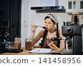 Young Asian female barista preparing fresh roasted coffee beans in a modern cafe kitchen with professional espresso equipment. 141458956