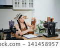 Young Asian female barista preparing fresh roasted coffee beans in a modern cafe kitchen with professional espresso equipment. 141458957