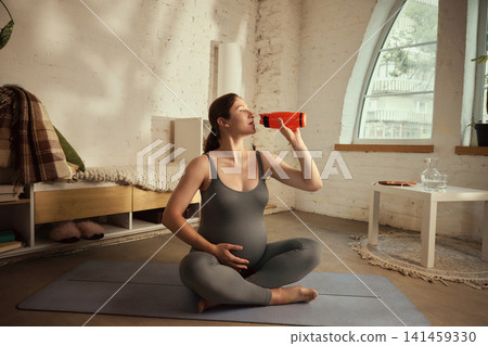 Thirsty young brunette pregnant woman drinking water after home workout 141459330