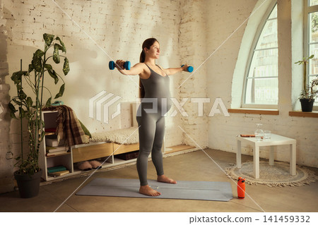 Active young expectant mother performing strength workout with dumbbells Active young expectant mother performing strength workout with dumbbells 141459332